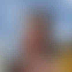 Close-up of the red clock tower at Pena Palace in Sintra, Portugal. The tower features four small turrets, a white clock face with Roman numerals, and Gothic-style detailing. Adjacent sections of the palace include a yellow domed structure and a black-and-white striped spire, all set against a cloudy sky.