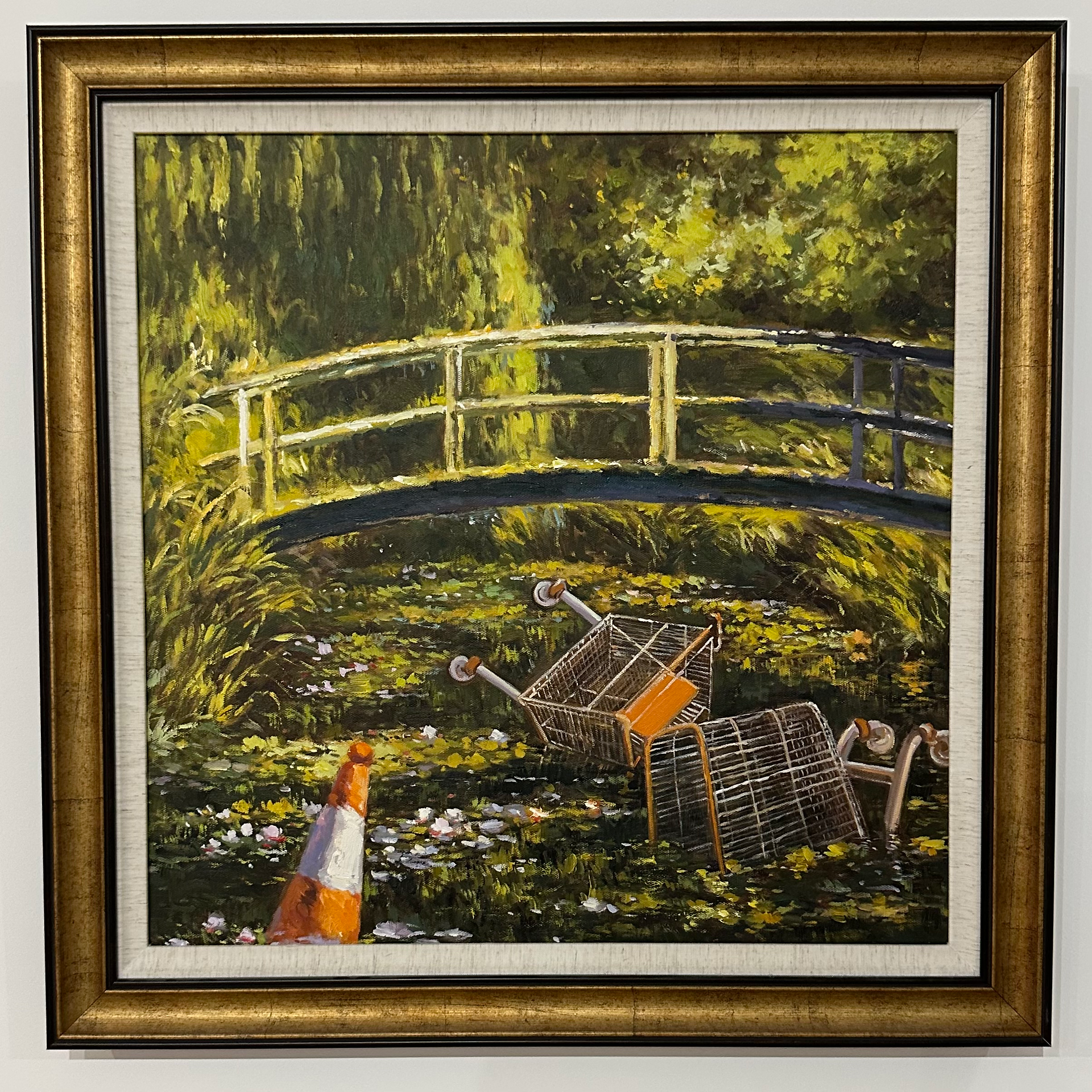 Serene pastoral scene featuring a small wooden bridge and dense greenery, disrupted by two overturned shopping carts and an orange traffic cone in the foreground. The contrast between nature and urban refuse evokes environmental critique.