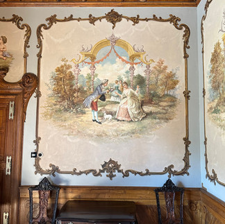 Painted wall inside the Regaleira Palace at Quinta da Regaleira in Sintra, Portugal. The mural shows a man in historical attire kneeling before a seated woman in a lush garden setting, framed by ornate decorative borders. Below the painting are two wooden chairs and a small table against the wall.