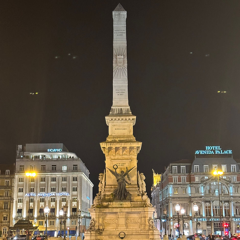Illuminated stone monument at night with sculpted figures, surrounded by lit buildings labeled "Rossio" and "Hotel Avenida Palace."