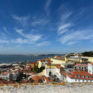 Panoramic view from the terrace of Lisbon’s National Pantheon, overlooking a sunlit cityscape of red-tiled rooftops and historic buildings. In the distance, the Tagus River stretches across the horizon, with the far shoreline visible under a clear blue sky. The vantage point captures the architectural charm and coastal beauty of Lisbon from above.