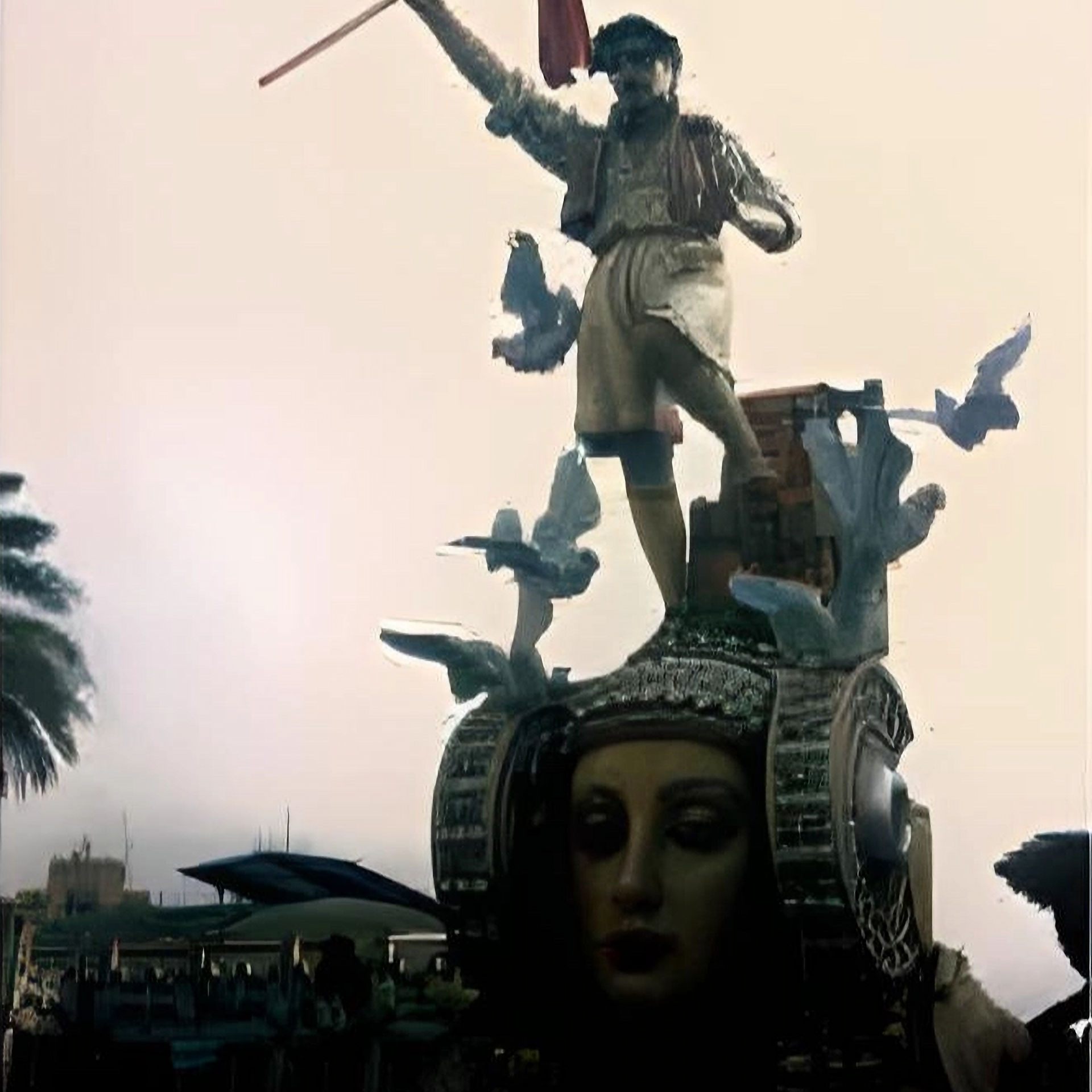Large, colorful falla sculpture displayed outdoors during Las Fallas festival in Valencia. The central figure is a stylized man with exaggerated features, wearing a dramatic costume and holding a flag. Sculpted birds appear to fly around him, adding movement and symbolism. The scene is framed by palm trees and surrounding buildings, capturing the festive and artistic spirit of the celebration.