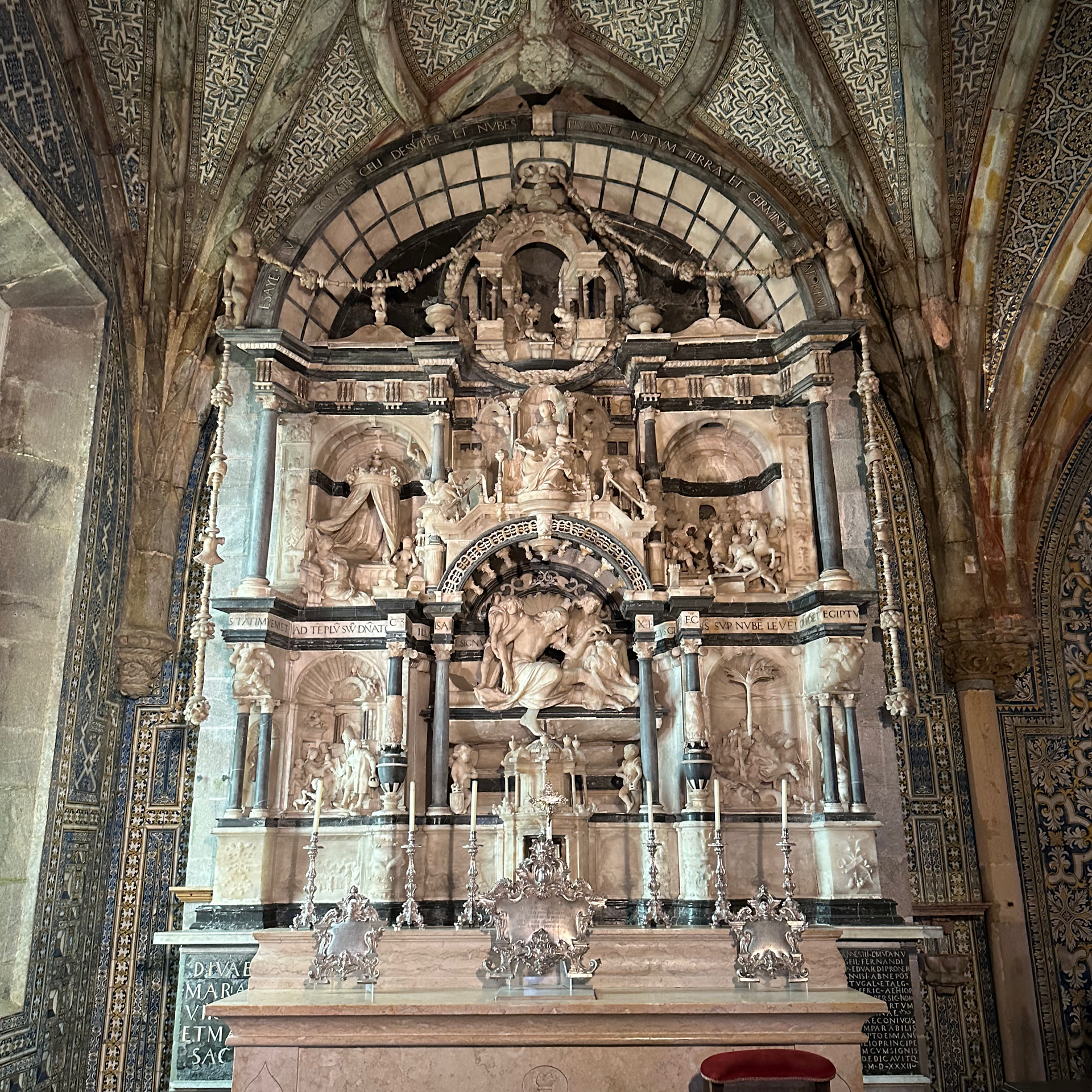 Ornate altar in the chapel at Pena Palace in Sintra, Portugal, carved from alabaster and limestone. The altarpiece features detailed reliefs depicting scenes from the life of Christ, framed by columns and decorative arches. The surrounding chapel interior includes vaulted ceilings and patterned tilework, reflecting a blend of Manueline and Renaissance styles.