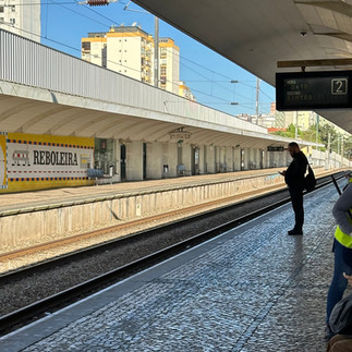Outdoor train platform at Reboleira station in Lisbon, Portugal. A white sign with bold black letters reads 'REBOLEIRA.' Passengers wait on the far platform beneath a long shelter. Train tracks run between the platforms, with residential buildings and a clear blue sky in the background.
