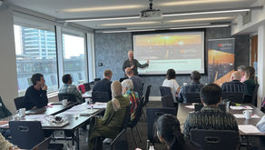 A man stands at the front of a room of people listening attentively. He gestures at a presentation which says 'SPRITE+ Innovations in Data Synthesis'