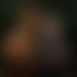 A woman with short white hair is standing outdoors next to a chestnut horse. She is smiling broadly and laughing, holding a colorful lead rope attached to the horse’s blue halter with a patterned noseband. The woman wears a dark green V-neck T-shirt and black pants, with a lanyard and whistle around her neck. The horse stands calmly beside her, ears forward, with a relaxed posture. Behind them are leafy trees and some farm structures, suggesting a rural setting. The moment feels natural, joyful, and connected.