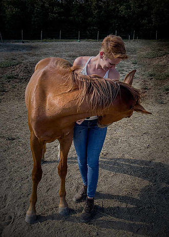 a woman with short red hair, jeans and tanktop, in a dry pasture with her chestnut young horse, who is bending around her to smell her butt :-)
