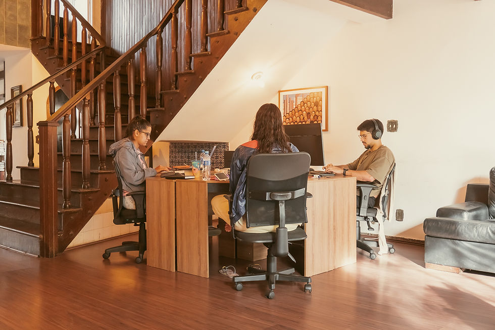 Three people work at a wooden desk in a sunlit home office under a staircase, with computers, headphones, and a calm focus.