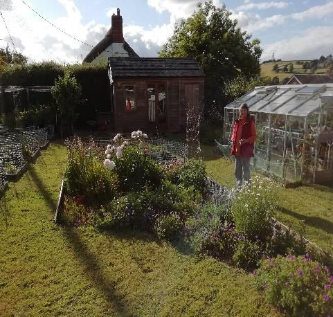 Colour photograph of a person in a red jacket standing in a lush garden with colourful flowers, next to a greenhouse and a wooden shed, under a partly cloudy sky.