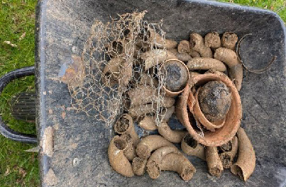 A wheelbarrow filled with old terracotta pots, wire mesh, and cow horns filled with biodynamic preparation which were dug up from being buried. It's on grass, suggesting outdoor garden work.