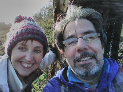 A smiling man and woman in winter clothing pose for a selfie outdoors by a tree, with sunlight filtering through. The mood is cheerful.