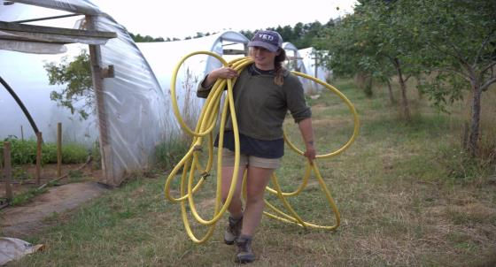 Person in casual attire carrying a coiled yellow hose on a farm, walking past polytunnels and trees, wearing a cap reading YETI.