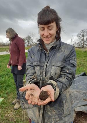 Woman in muddy jacket smiling, holding soil in hands. Another person in background on grassy field under cloudy sky. Rural setting.