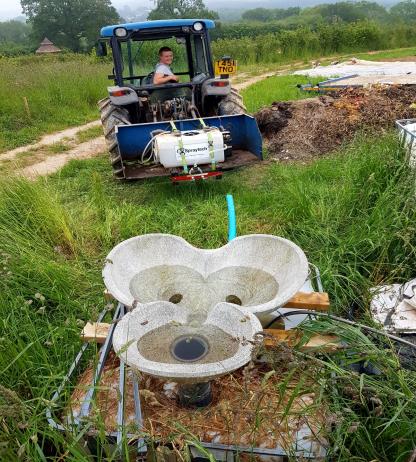 A person drives a blue tractor on a grassy path. In the foreground, there's a concrete structure with bowls. Lush greenery surrounds the area.