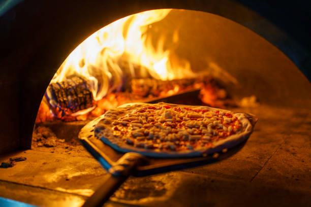 Pizza on a wooden peel in a wood fired brick oven with flames. The setting is warm and rustic, with fire casting an inviting glow.