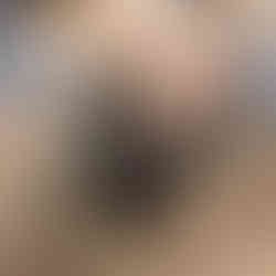 Young boy in a blue hat leans over a black bucket, hand submerged. Seated adults nearby, outdoors.