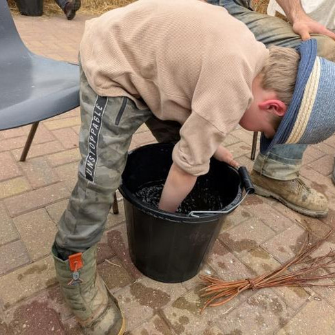 Young boy in a blue hat leans over a black bucket, hand submerged. Seated adults nearby, outdoors.