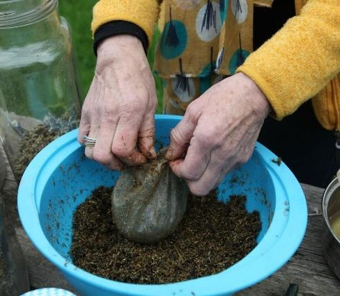 Woman wearing a yellow sweater, her hands tying up a stag bladder over a blue bowl on a wooden table, surrounded by jars and green grass in the background.