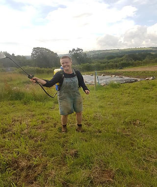 A smiling person wearing overalls holding out a long sprayer in a grassy field. A blue sky, rolling hills and trees are in the background.