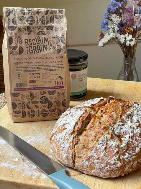 Rustic bread loaf on a cutting board with a serrated knife, flour bag labeled "Reclaim the Grain," and a jar of blackcurrant jam. Dry flowers in a small vase in the background.