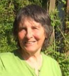 Smiling woman in a bright green shirt stands outdoors with lush greenery in the sunny background, creating a cheerful and natural atmosphere.