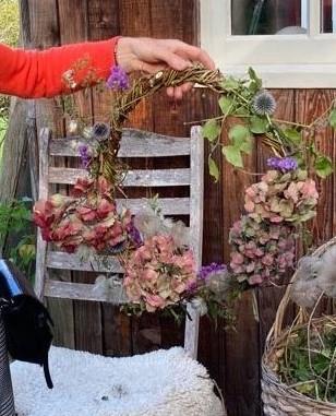 Person holding a circular floral wreath of pink and purple flowers and green leaves. Rustic wooden chair and basket in the cozy background.
