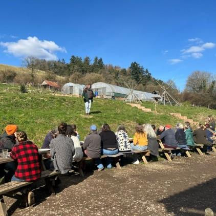 A group of people sitting at picnic tables in a long row under a clear blue sky, listening to a speaker in a rural setting with polytunnels and trees in the background.