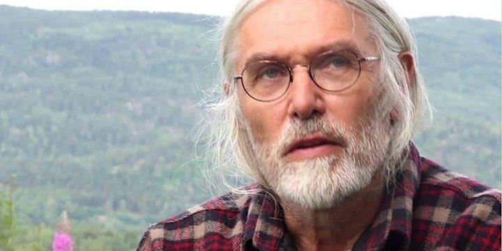 Elderly man with glasses and long gray hair in a checkered shirt, looks thoughtful against a hilly, green landscape background.