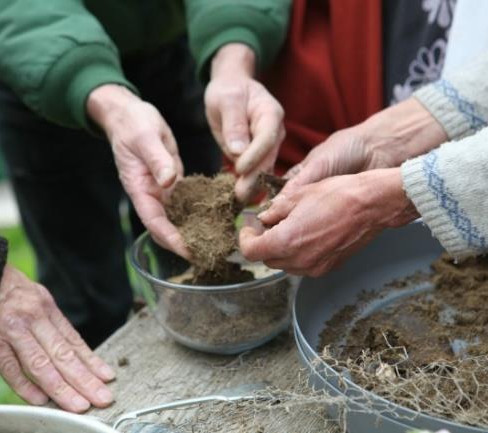 Hands sorting biodynamic preparations over a glass bowl outdoors, with green and brown sleeves of clothing visible. The setting is natural and collaborative.