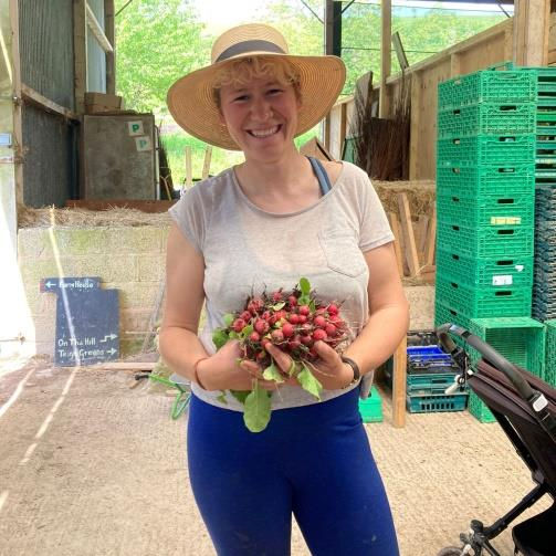 Smiling woman in a straw hat holds radishes inside a greenhouse. Green crates are stacked behind. Warm, sunny ambiance.