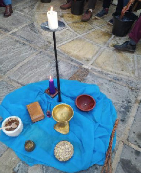 Candles and ritual items on a blue cloth on a stone floor. People seated around in the background. A lit white candle stands on a pole, creating a serene mood.