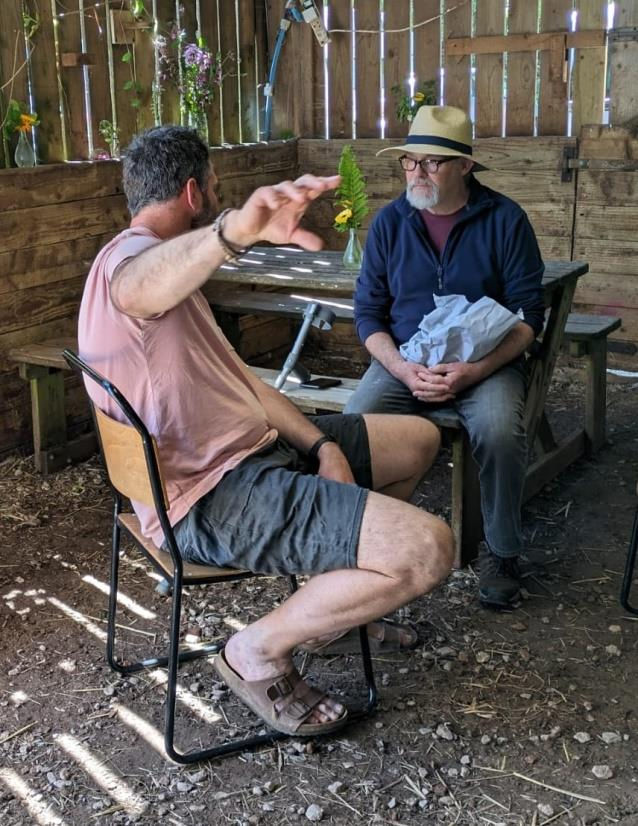 Two men sit in a wooden, rustic setting; one sitting on a chair gestures while talking. The other man wearing a hat sitting on a picnic table seat listens, holding a bag. Flowers in a vase are on a wooden picnic table.
