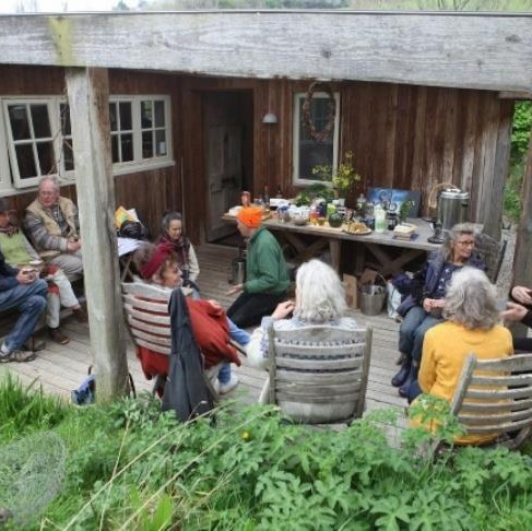 A group of ten people sit on chairs on a wooden outdoor porch chatting in front of a wooden table. The atmosphere is relaxed with greenery in the background.