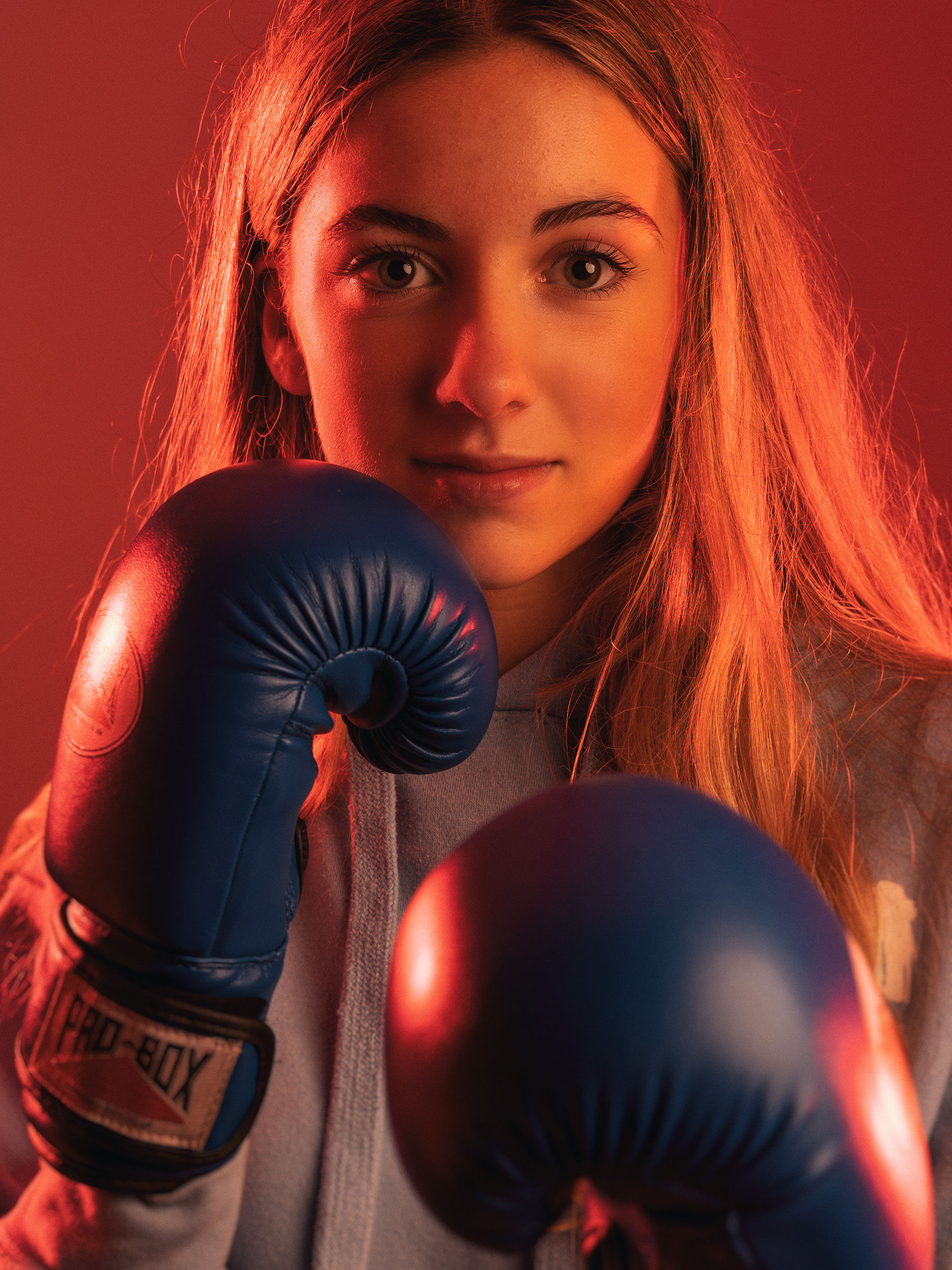 Teenage Girl with dramatic orange lighting as she looks straight to camera with blue boxing gloves poised around her facec