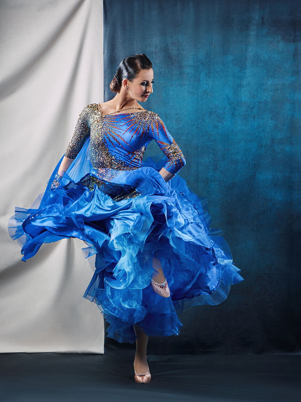 Female Ballroom dancer in a stunning blue dress simply lifting for a kick as she gazes with a smile to the right