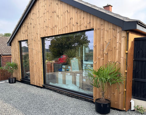 Modern wooden clad building with large windows, gravel path, potted plants.