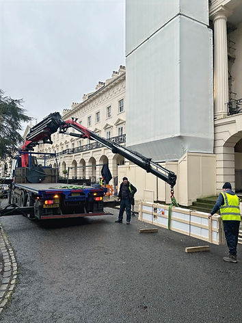 Crane lifting large white structure, workers on site, exterior of building in background.
