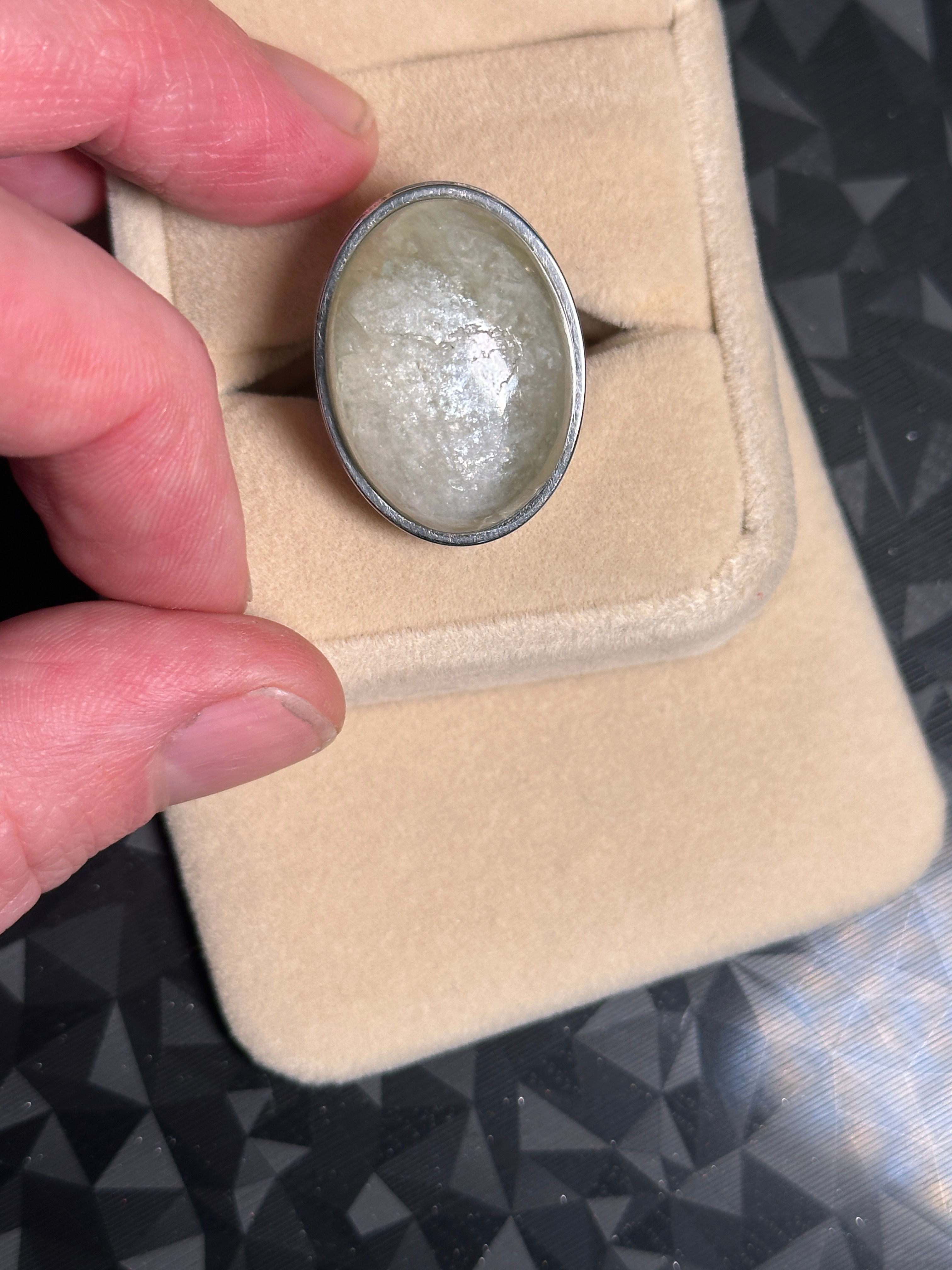 Bague ovale avec pierre claire tenue par une main sur support beige.