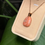 Miniature : Pendentif orange sur chaîne dorée, présenté dans un écrin beige, devant une plante.
