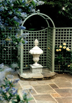 a statue of a vase with a pine cone on top is in a garden surrounded by flowers trellis and structures .