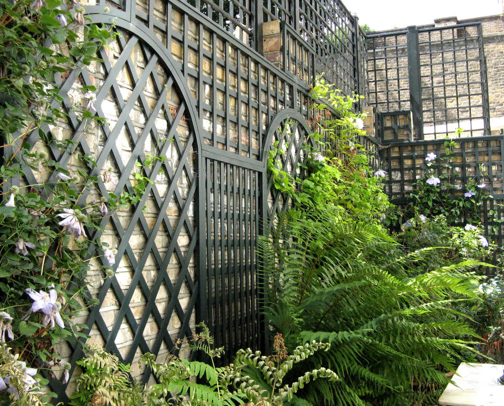 a brick wall with a dark green trellis surrounded by ferns and flowers