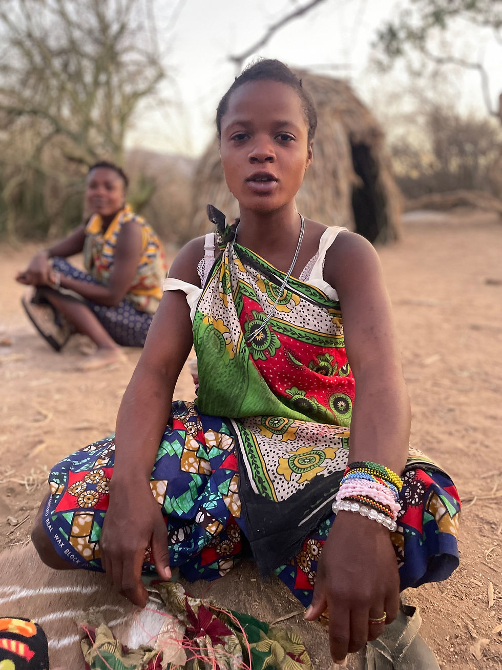 Hadzabe woman kneeling in the landscape near Lake Eyasi in Tanzania, with another woman standing behind her in a traditional Hadza camp.