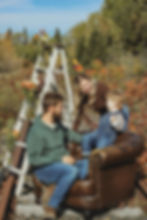 Family of three outdoors on a rustic chair by railroad tracks, smiling and interacting, with autumn foliage