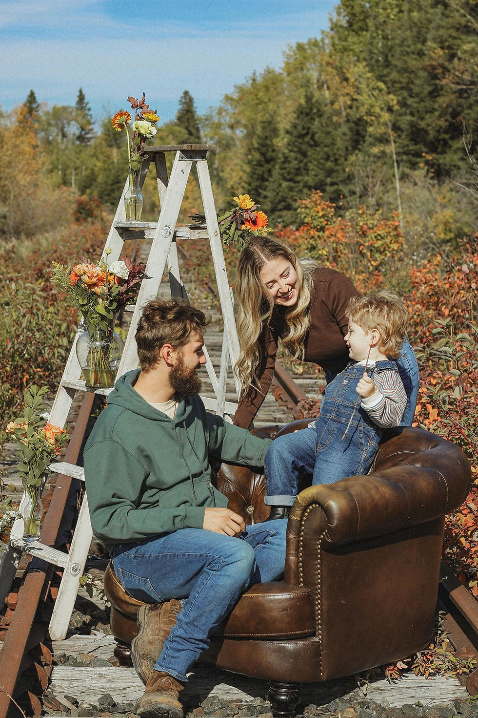 Family of three outdoors on a rustic chair by railroad tracks, smiling and interacting, with autumn foliage