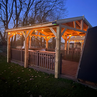 A hot tub is tucked under the cover of a wooden gazebo lit up with festoon lights at night, there is a cosy warm orange glow!