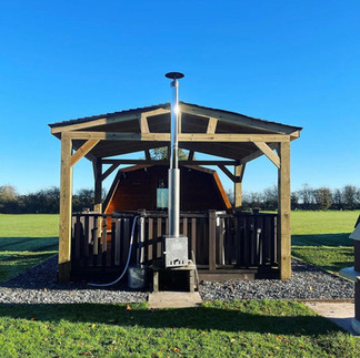 Looking out over decking with trees and a white brick oven-style BBQ in back ground. A picnic bencGlamping pod in lush green field. The hot tub sites on a deck, under the cover of a wooden gazebo