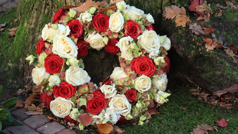 runder Blumenkranz aus weissen und roten Rosen für eine Beerdigung angelehnt an einen moosbewachsenen Baumstamm und umgeben von Herbstlaub