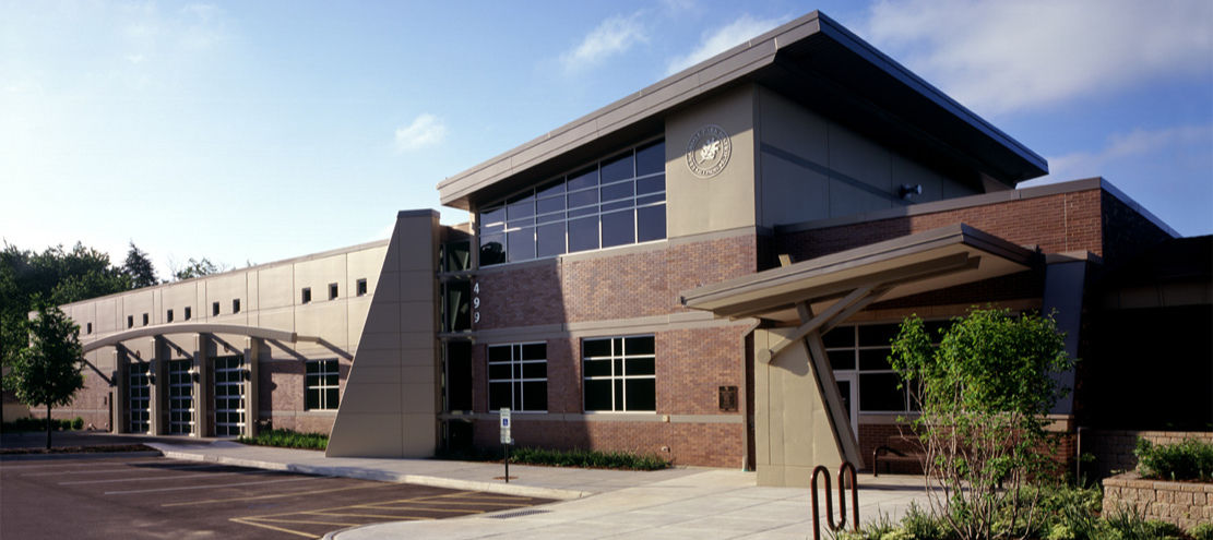 Exterior view of a public works facility with brick-and-metal architecture, large windows, covered entrance, and landscaped surroundings.