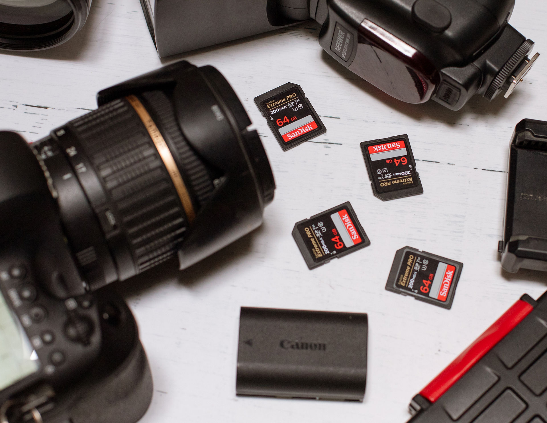 Close-up detail of Canon DSLR camera, flash, and SD cards spread out on a white tabletop, showcasing essential tools used by professional photographers.