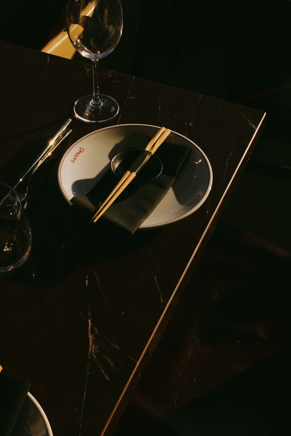 Overhead view of a restaurant place setting on dark marble - branded white plate, black napkin, wooden chopsticks and wine glass lit by a shaft of warm light